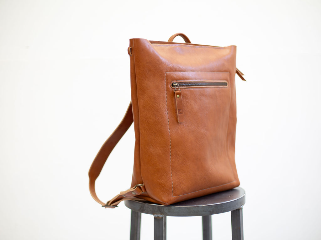 The convertible leather backpack is seen from an angle. The attachment points for the removable backpack straps and the front pocket with zipper are visible. 
