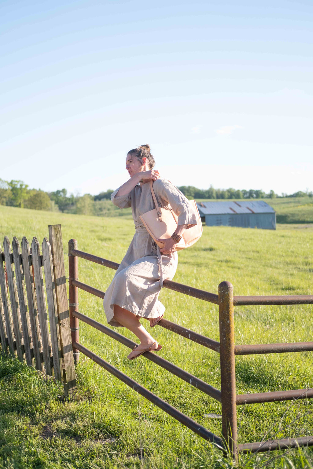 A woman sitting on a fence is getting ready to jump of a rustic fence, putting the large leather tote bag over her shoulder. 
