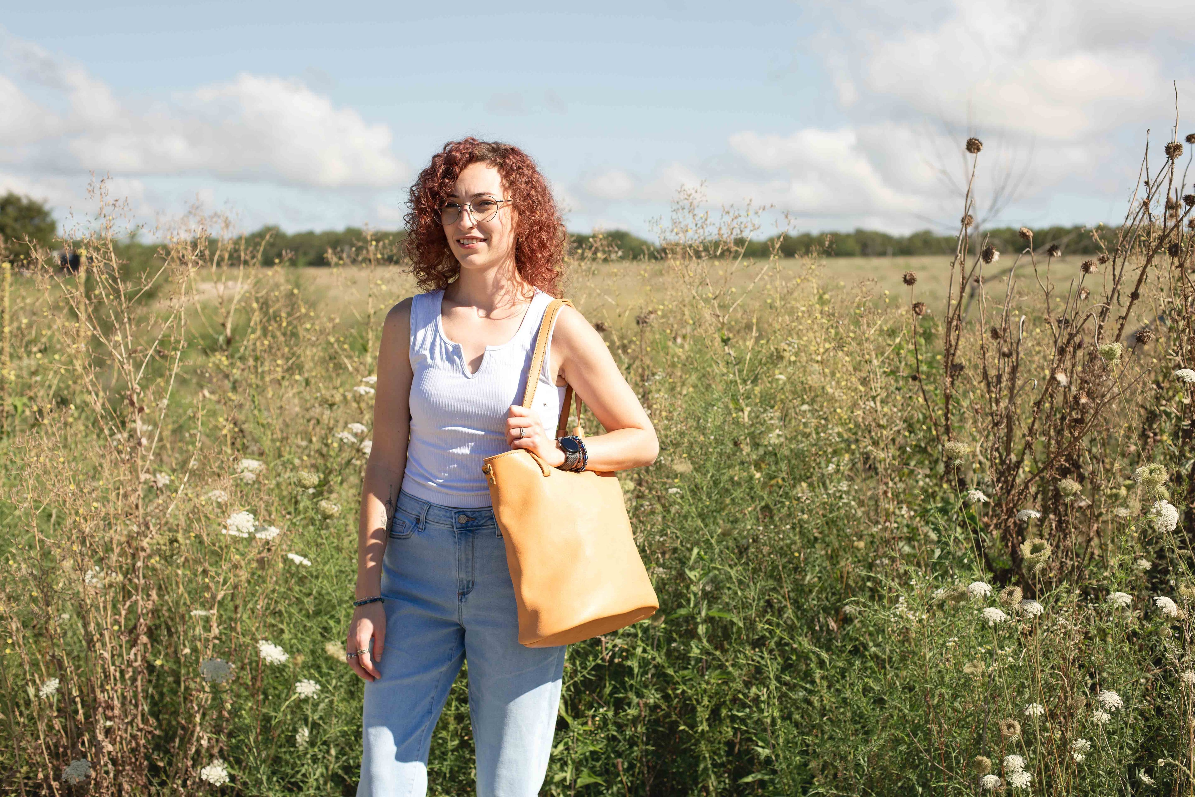 The large tan tote bag, model "The Gatherer" is shown on a model in a flower field. 