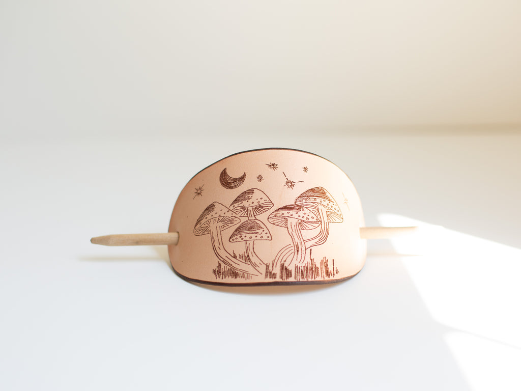 The leather hair clip shows an original art drawing of mushrooms at night that is laser engraved into the non-toxic, organic leather, sustainably sourced. 