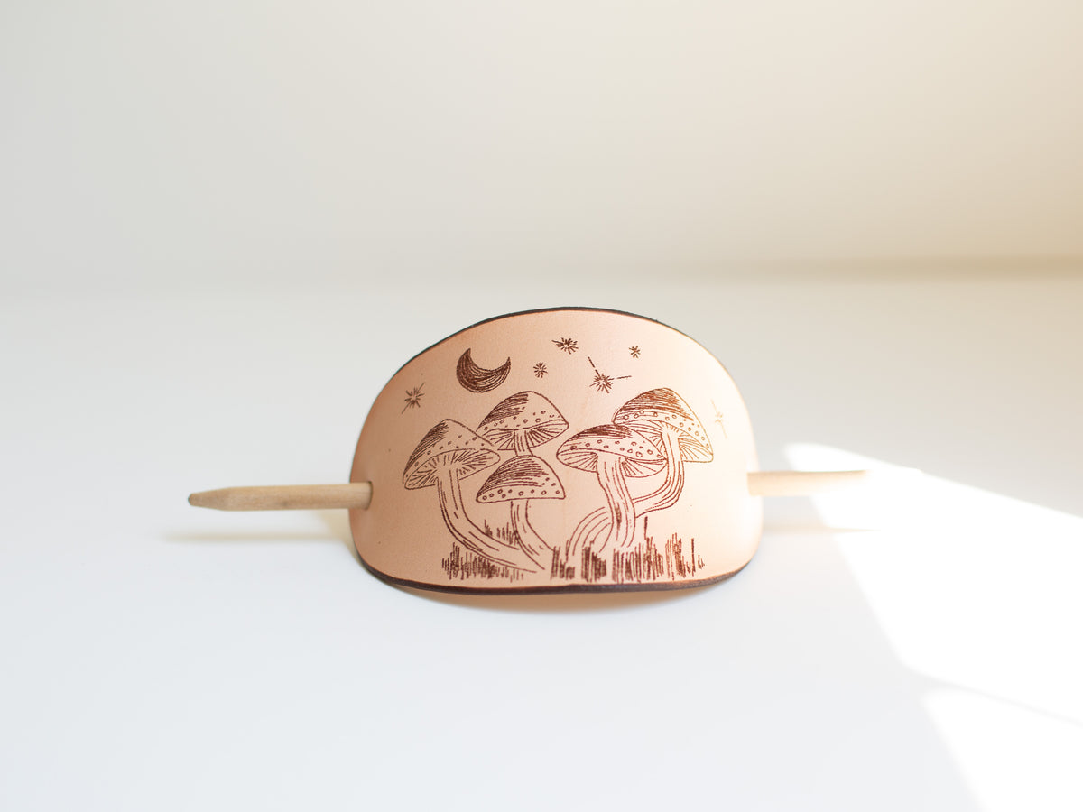The leather hair clip shows an original art drawing of mushrooms at night that is laser engraved into the non-toxic, organic leather, sustainably sourced. 