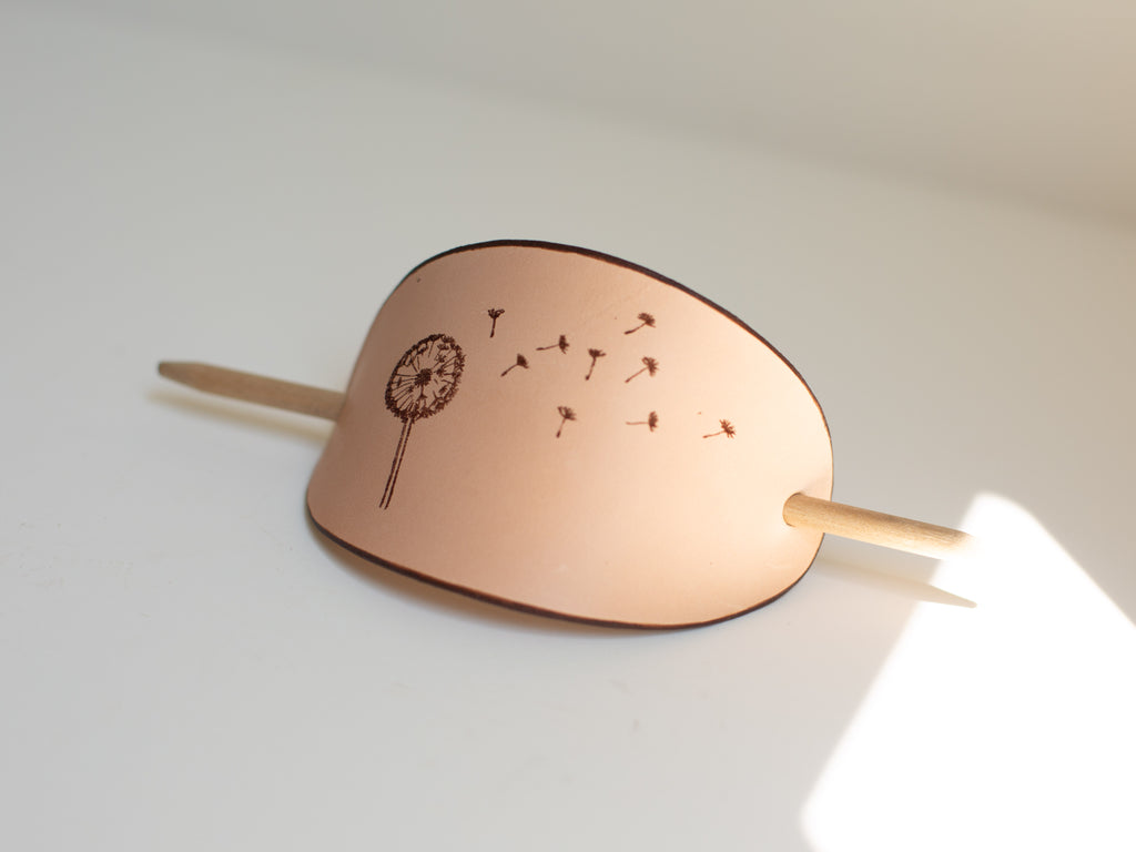 The seed heads from the laser engraved dandelion are flying over the large leather barrette with wooden stick.
