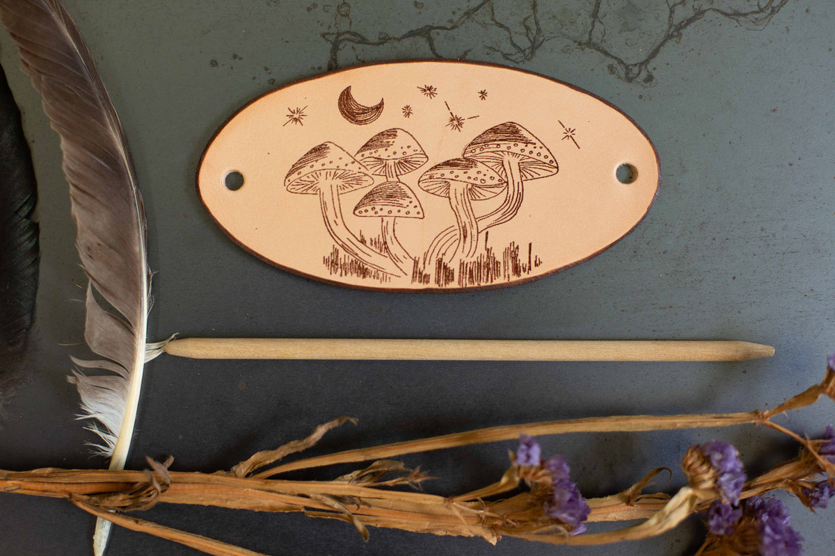 The large leather barrette is laying flat on a dark surface so that the whole laser engraved mushroom drawing is visible. 