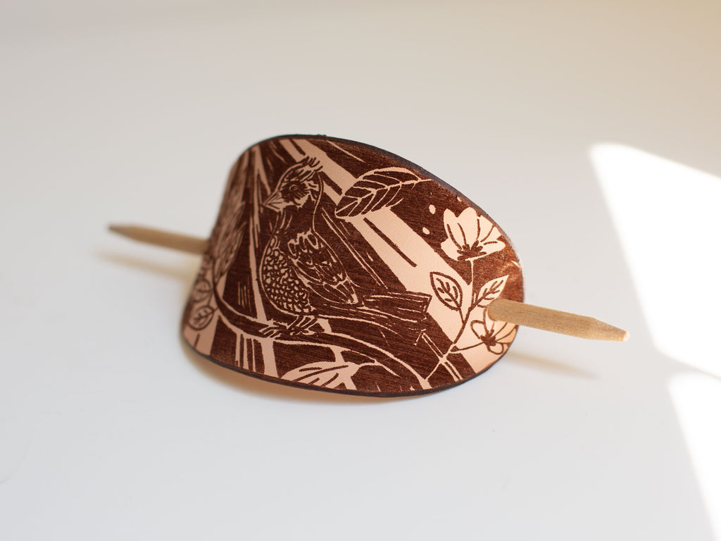 The leather hair slide has a laser engraved artistic drawing of a bird sitting on a branch in copper cut linocut style. 