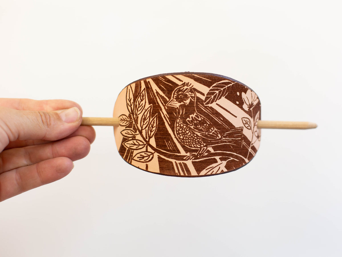 The large leather barrette is made with organic leather and has a laser engraving that shows the artistic rendering of a bird in a forest with sunlight streaming in. 
