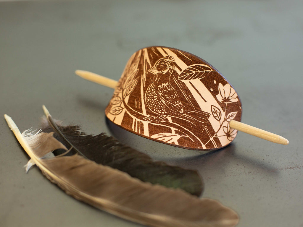 The large leather barrette has a laser engraved original art drawing in linocut style that shows a little bird on a branch with sunlight streaming in. 